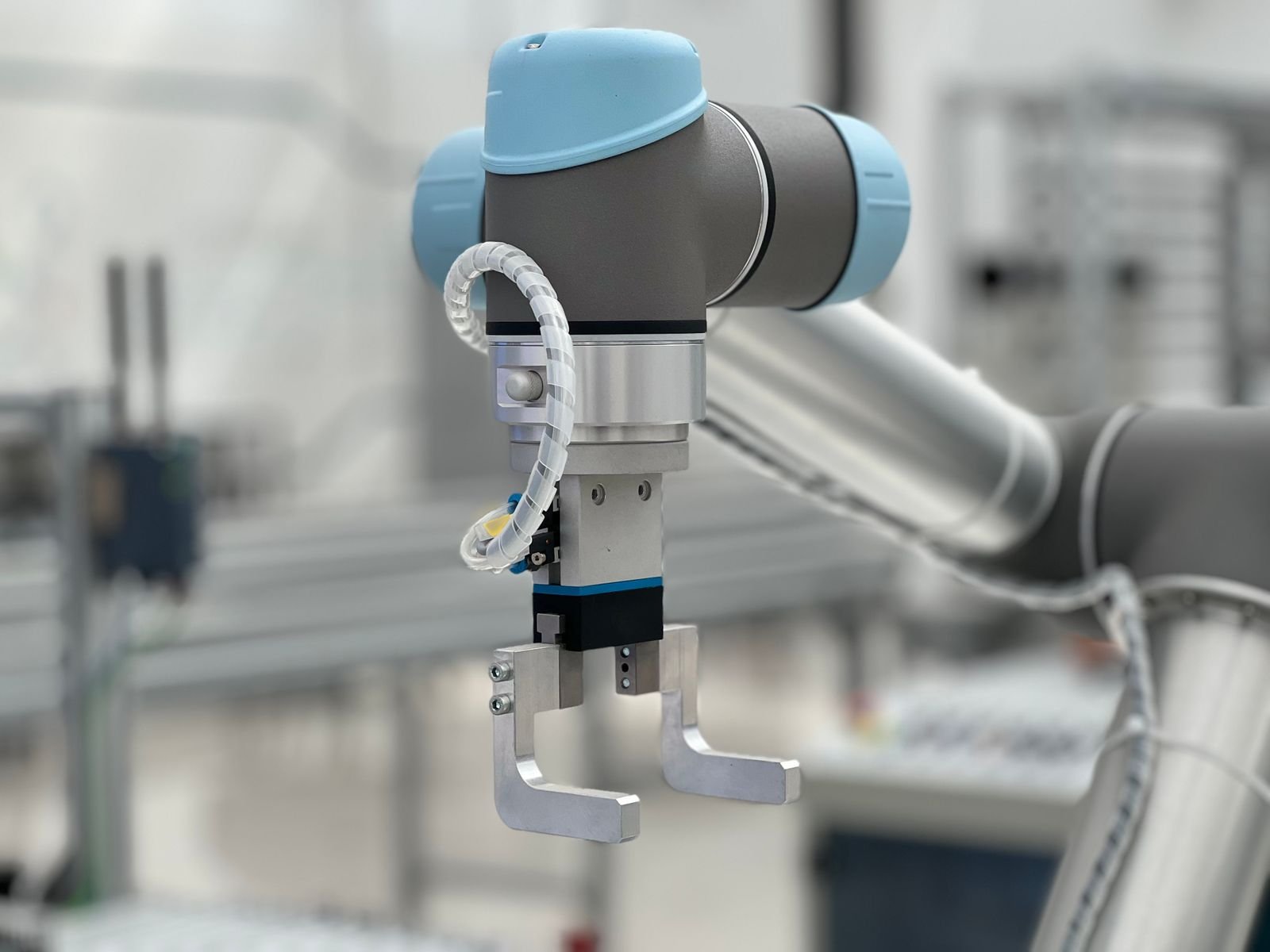 Garra de braço robótico industrial em close, com cabeça azul clara e duas pinças metálicas, sobre fundo de fábrica desfocado