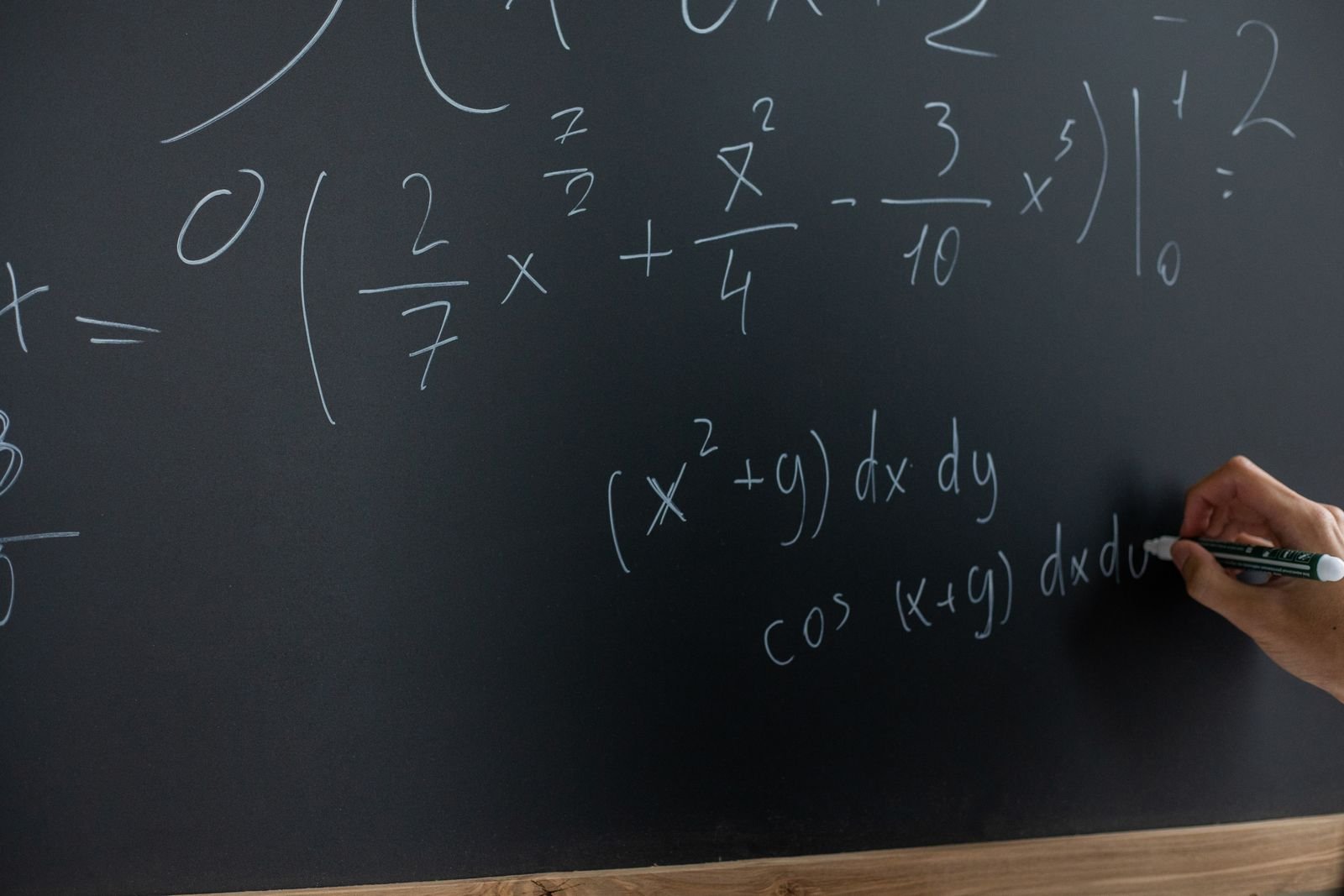 Quadro preto com equações de cálculo escritas à mão em giz branco, e uma mão escrevendo à direita